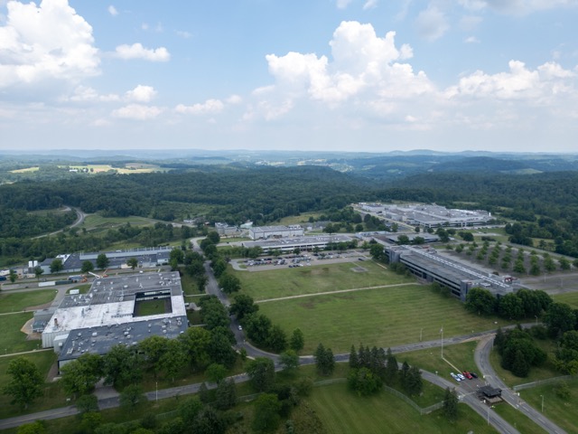 Aerial view of a large complex of buildings surrounded by parking lots, roads, and green fields, with forested hills in the background under a partly cloudy sky.