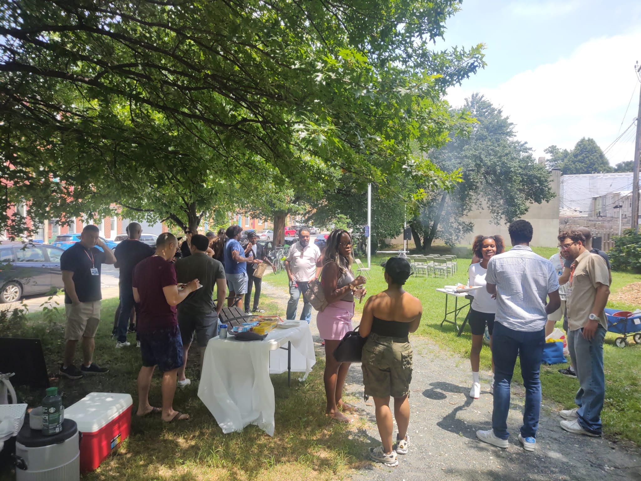 People gather outdoors under trees, standing and talking near tables with food and drinks at a casual daytime event.