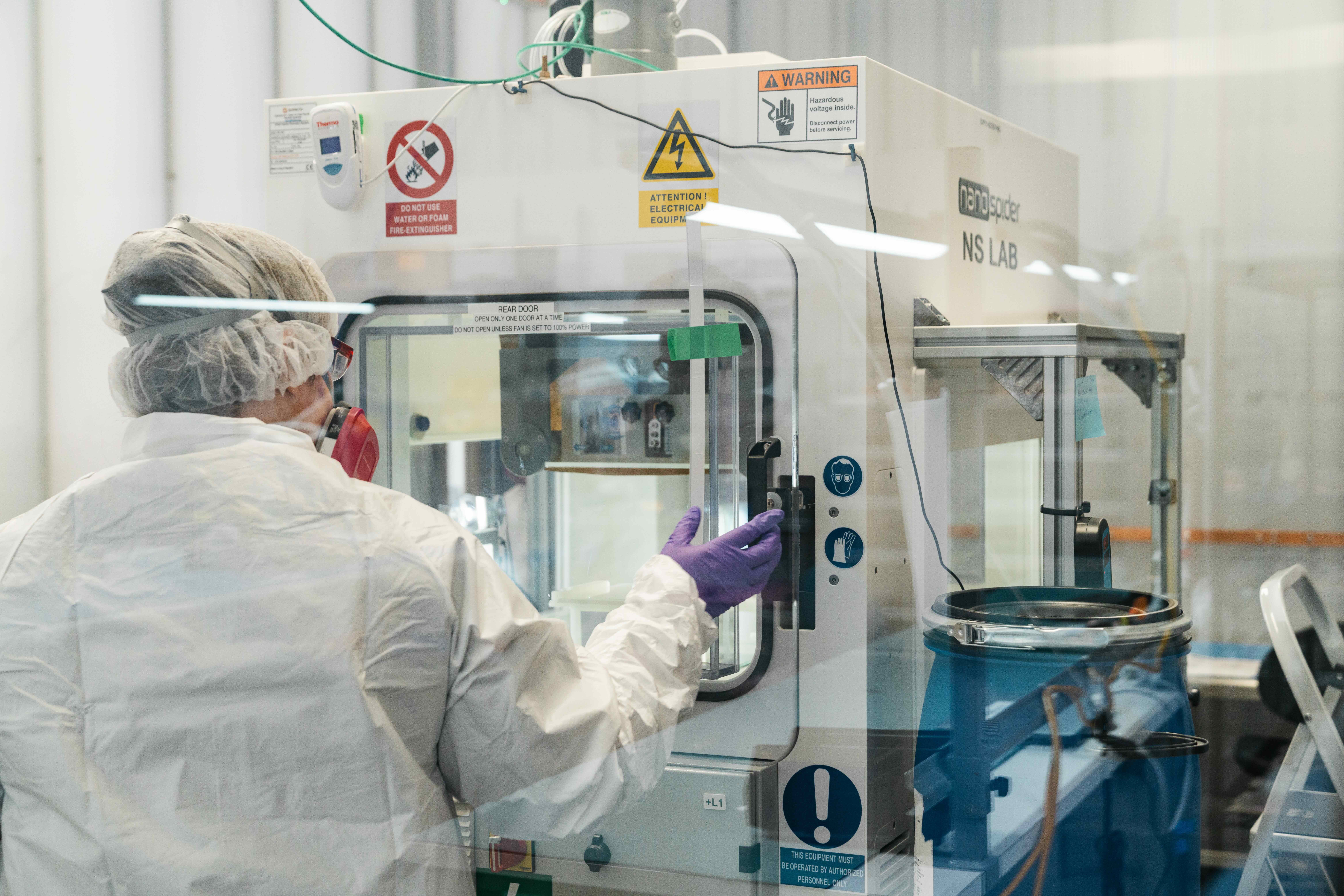 A person in protective gear operates laboratory equipment inside a sterile lab environment, with warning signs and safety notices visible on the machine.