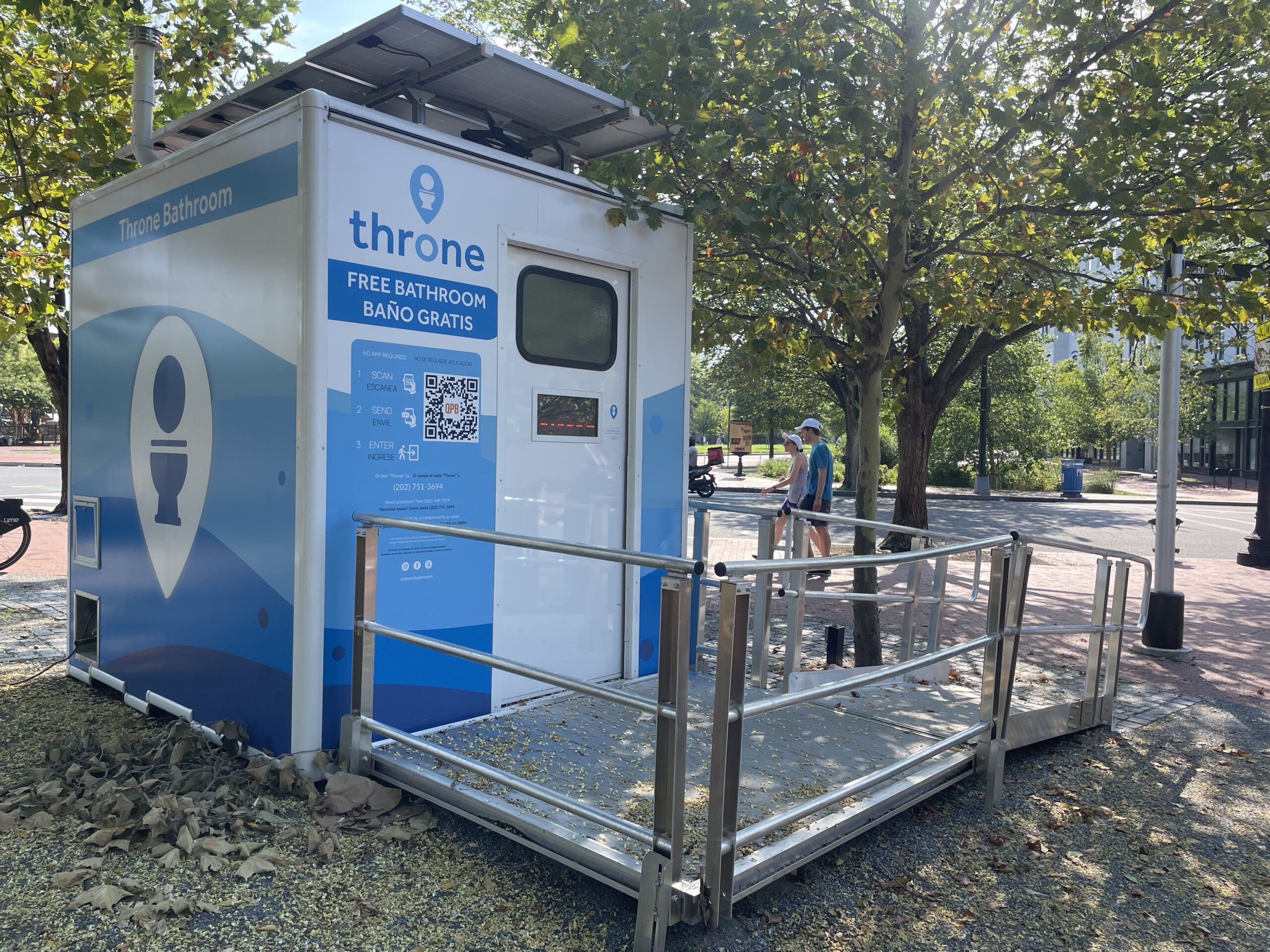 A standalone public restroom unit labeled "throne bathroom" with a ramp, solar panel, and signage offering free bathroom access, situated outdoors near trees and a sidewalk.