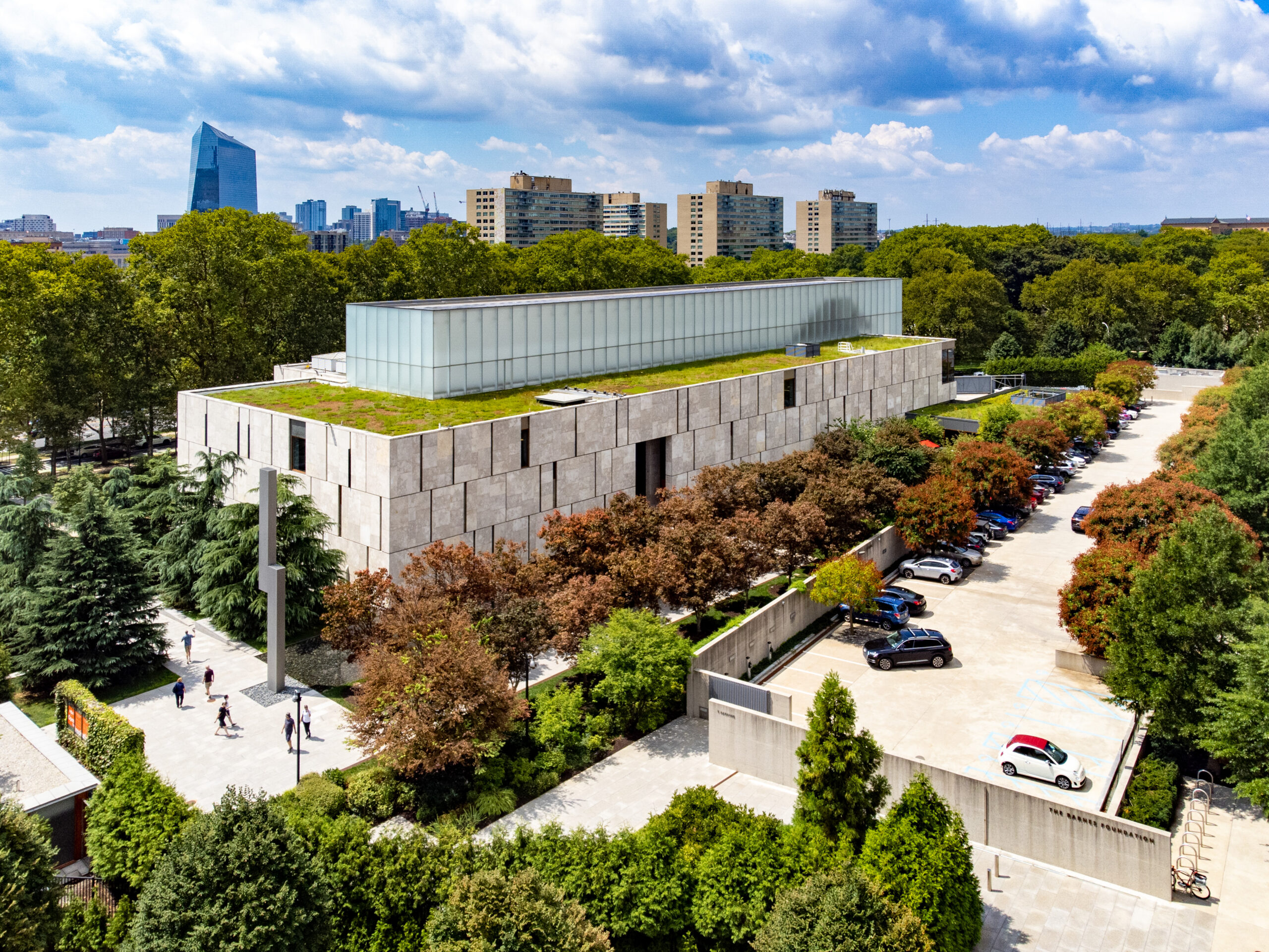 Aerial view of a modern rectangular building with a green roof, adjacent parking lot, trees, and a city skyline in the background under a partly cloudy sky.