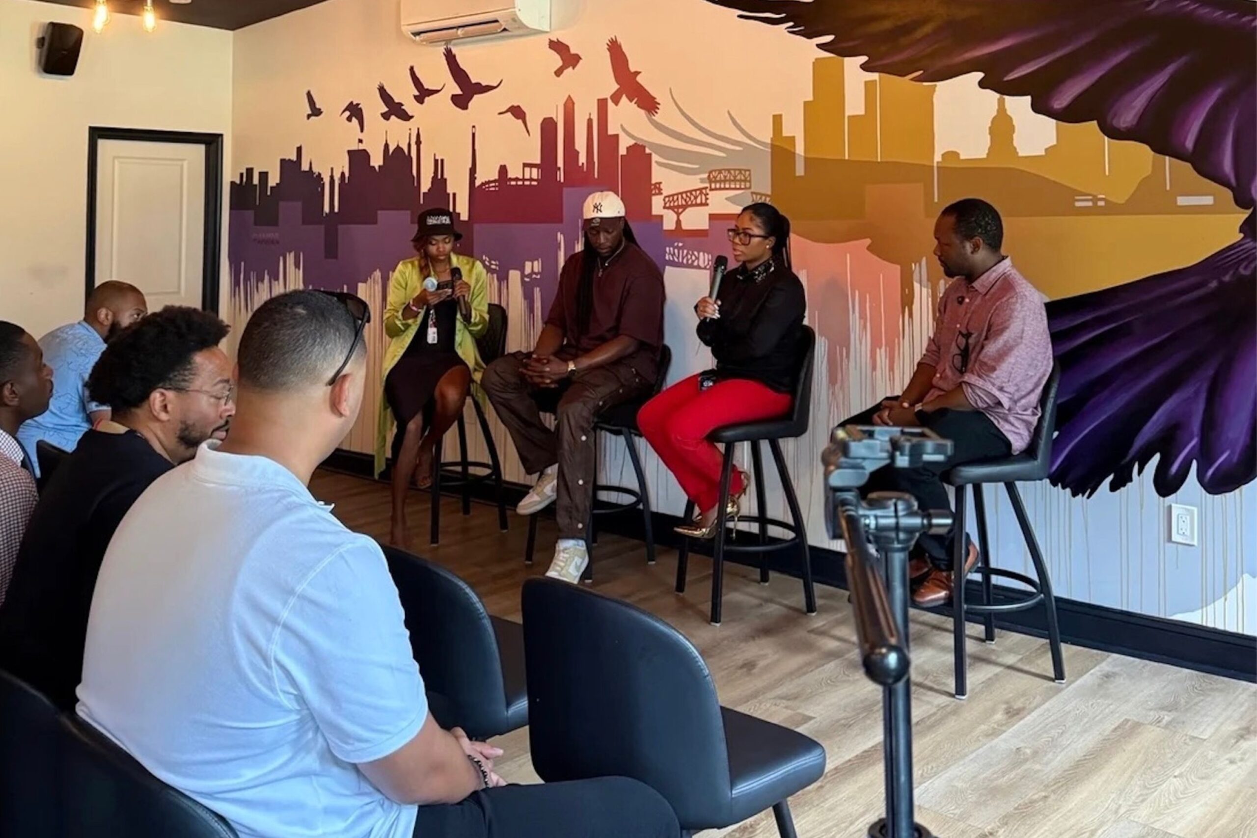 Four panelists speak to an audience in a small room with a city skyline mural. A camera on a tripod records the discussion.