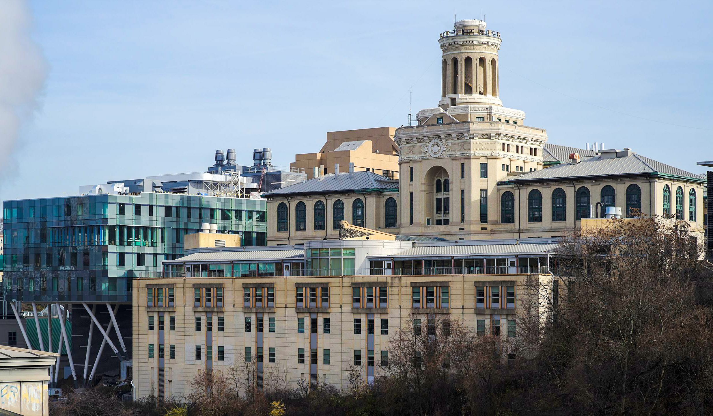 A large beige historic building with a central tower stands next to a modern glass structure under a clear sky.