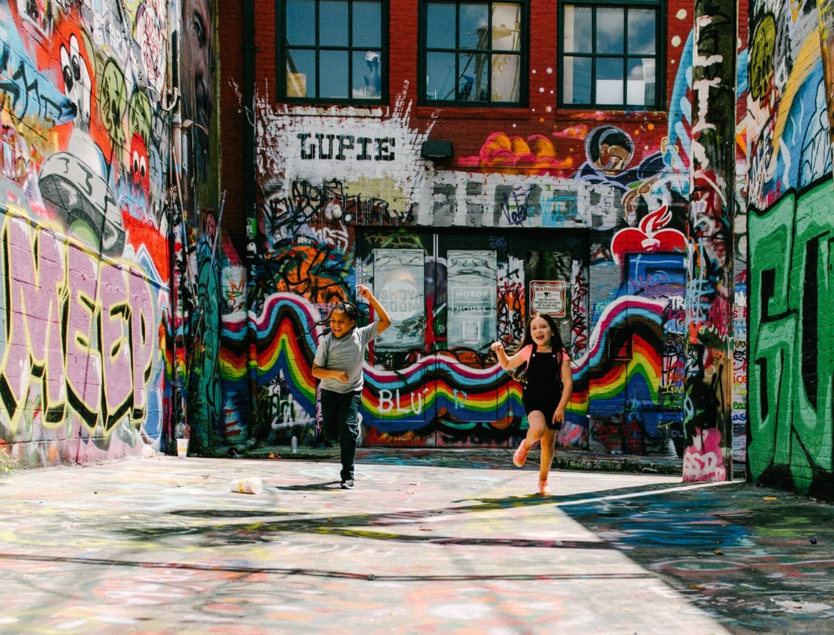 Two children run and play in a graffiti-covered alley on a sunny day, surrounded by colorful street art and murals.