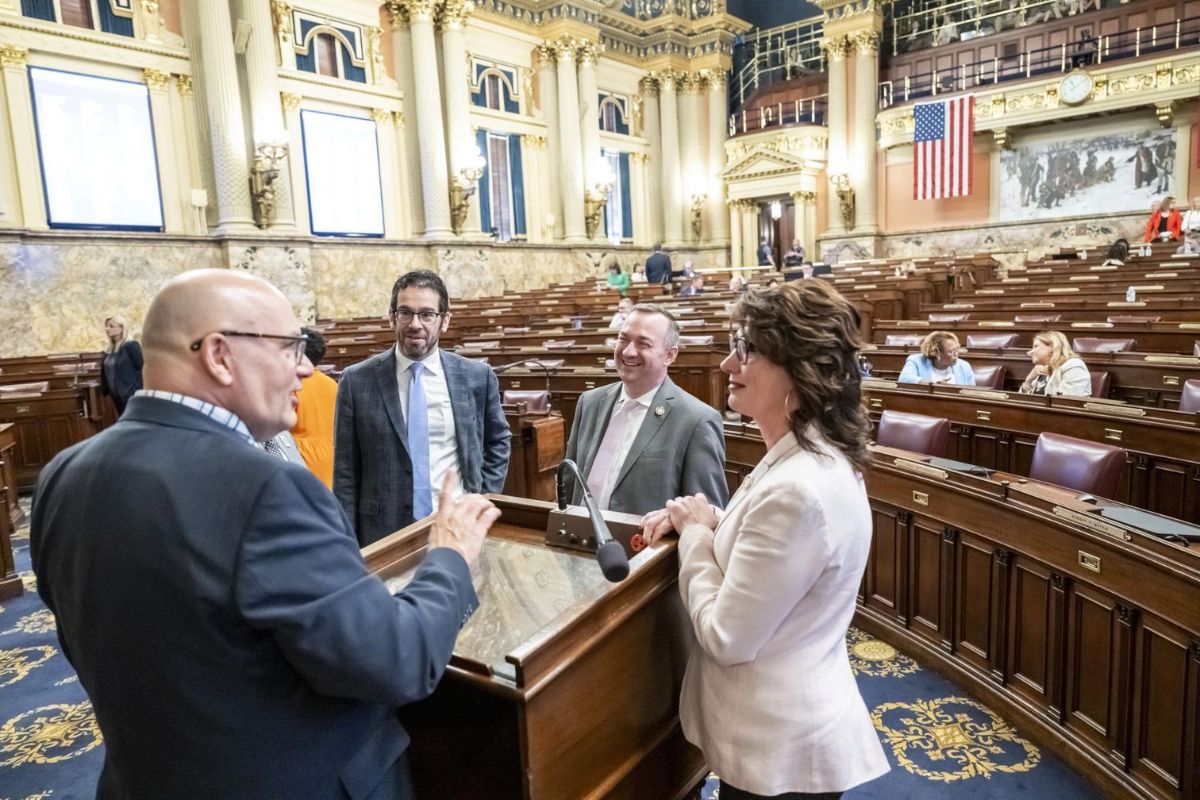 Four people stand and converse near a podium inside a large, ornate legislative chamber with desks, an American flag, and others seated in the background.