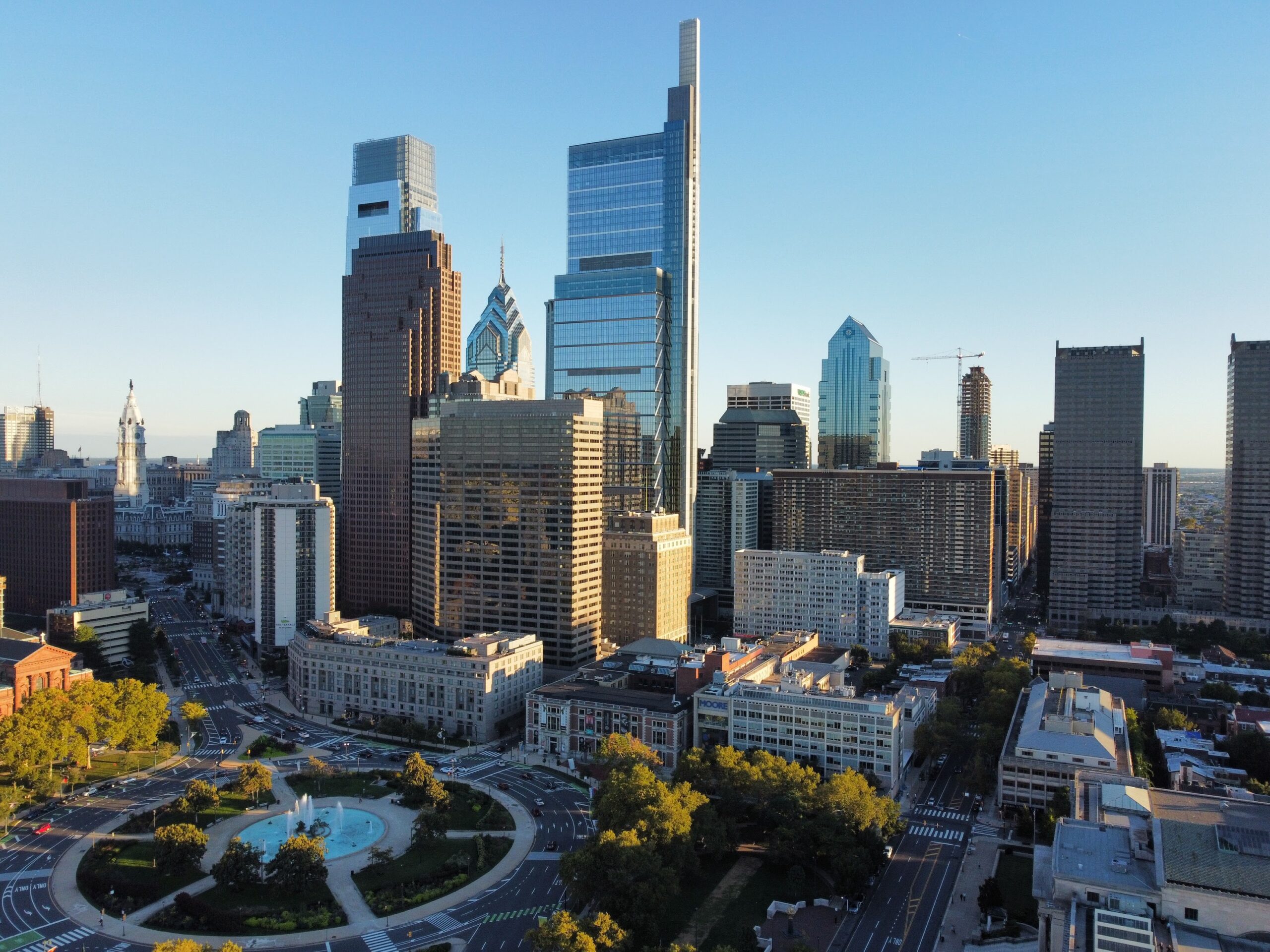 Aerial view of downtown Philadelphia showing modern skyscrapers, historic buildings, and a traffic roundabout with a central fountain.