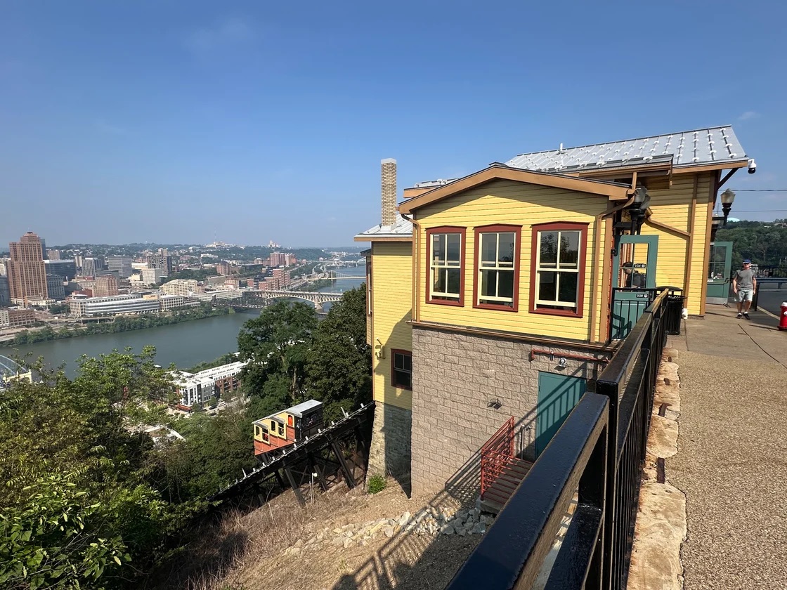 A yellow building sits atop a hill overlooking a city skyline and river, with a funicular railway descending the steep slope beside it.