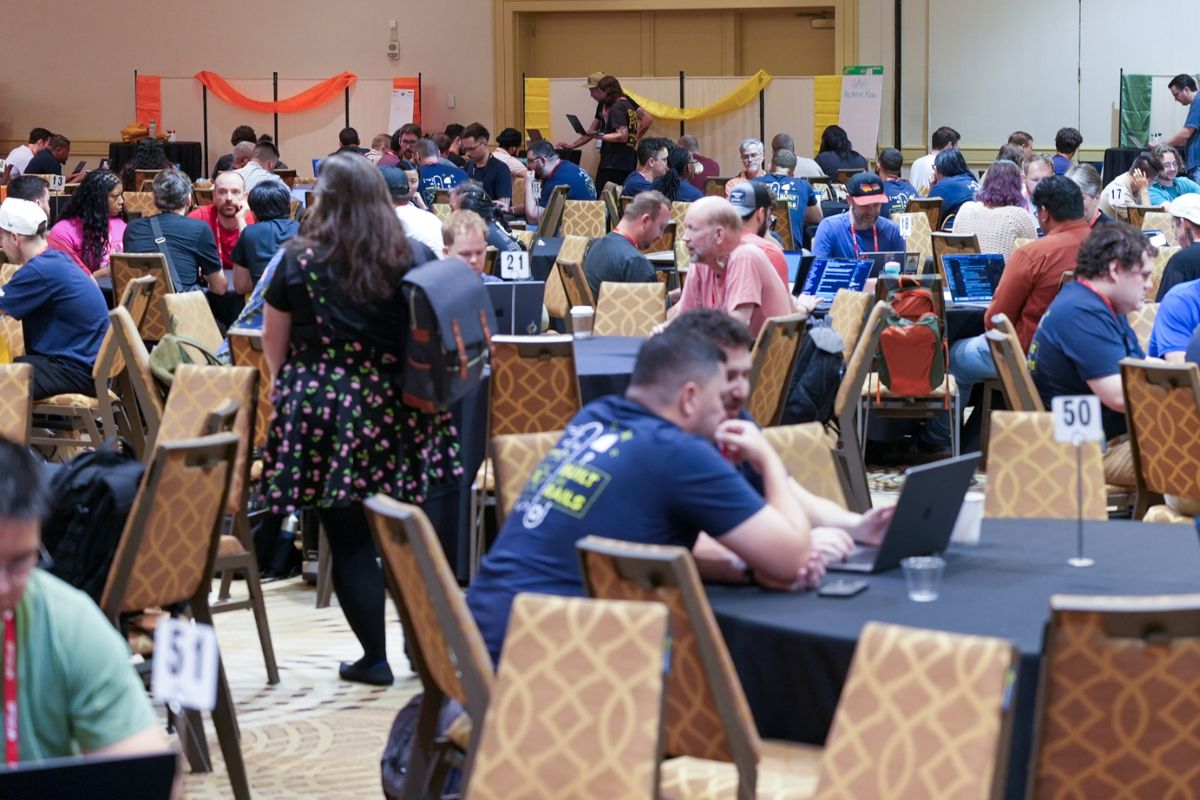 Large room with many people seated at round tables working on laptops; some are standing and talking, suggesting a conference or collaborative event.