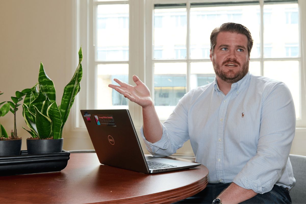 A man sits at a table with a laptop, gesturing with one hand. Two potted plants are on the table, and large windows are in the background.