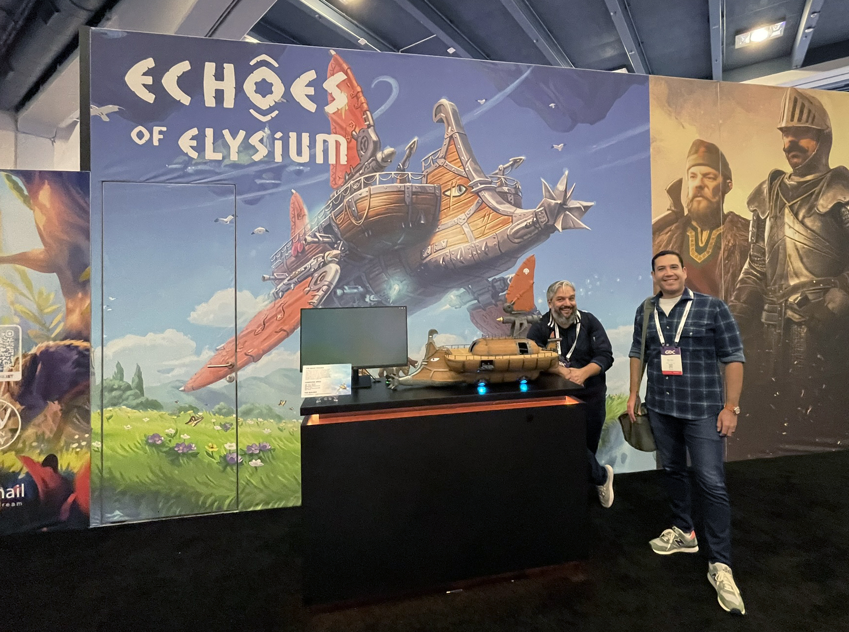 Two men stand by a display booth for “Echoes of Elysium” featuring a fantasy airship backdrop, a monitor, and a model airship on a black counter.