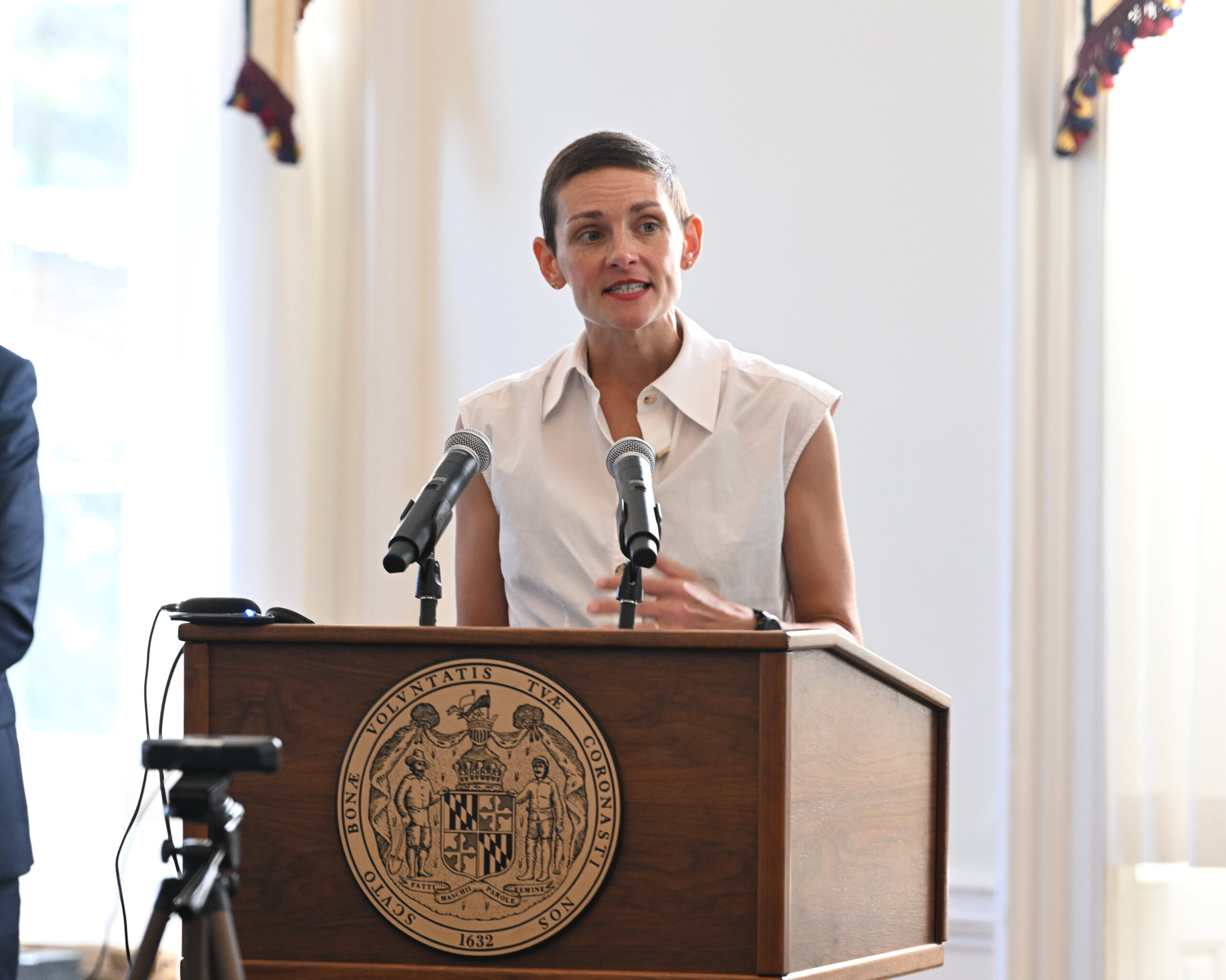 A person in a sleeveless white shirt speaks at a podium with two microphones and the Maryland state seal in a well-lit room.
