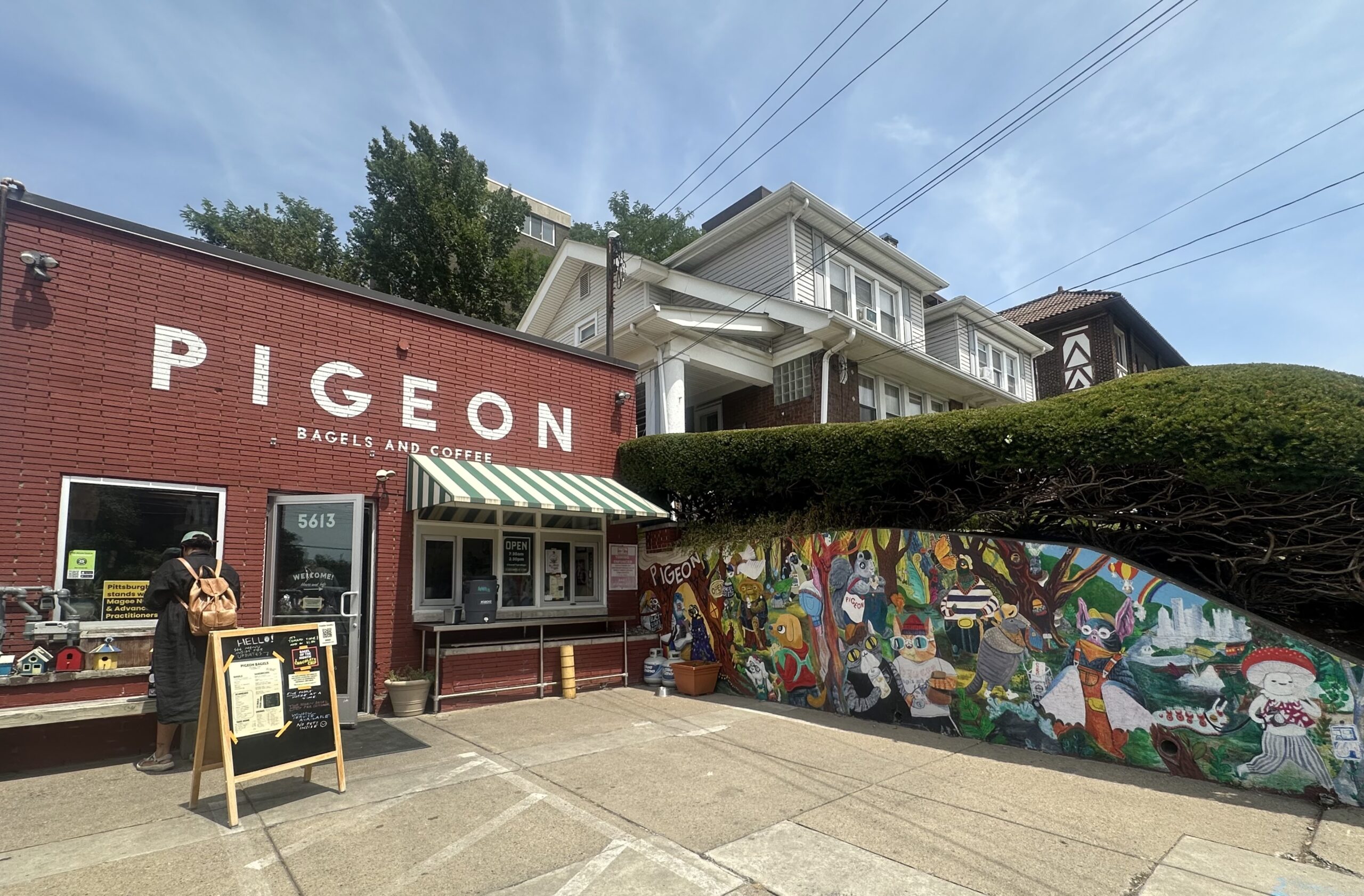 A red brick cafe called Pigeon Bagels and Coffee sits next to a colorful mural and a residential building on a sunny day.