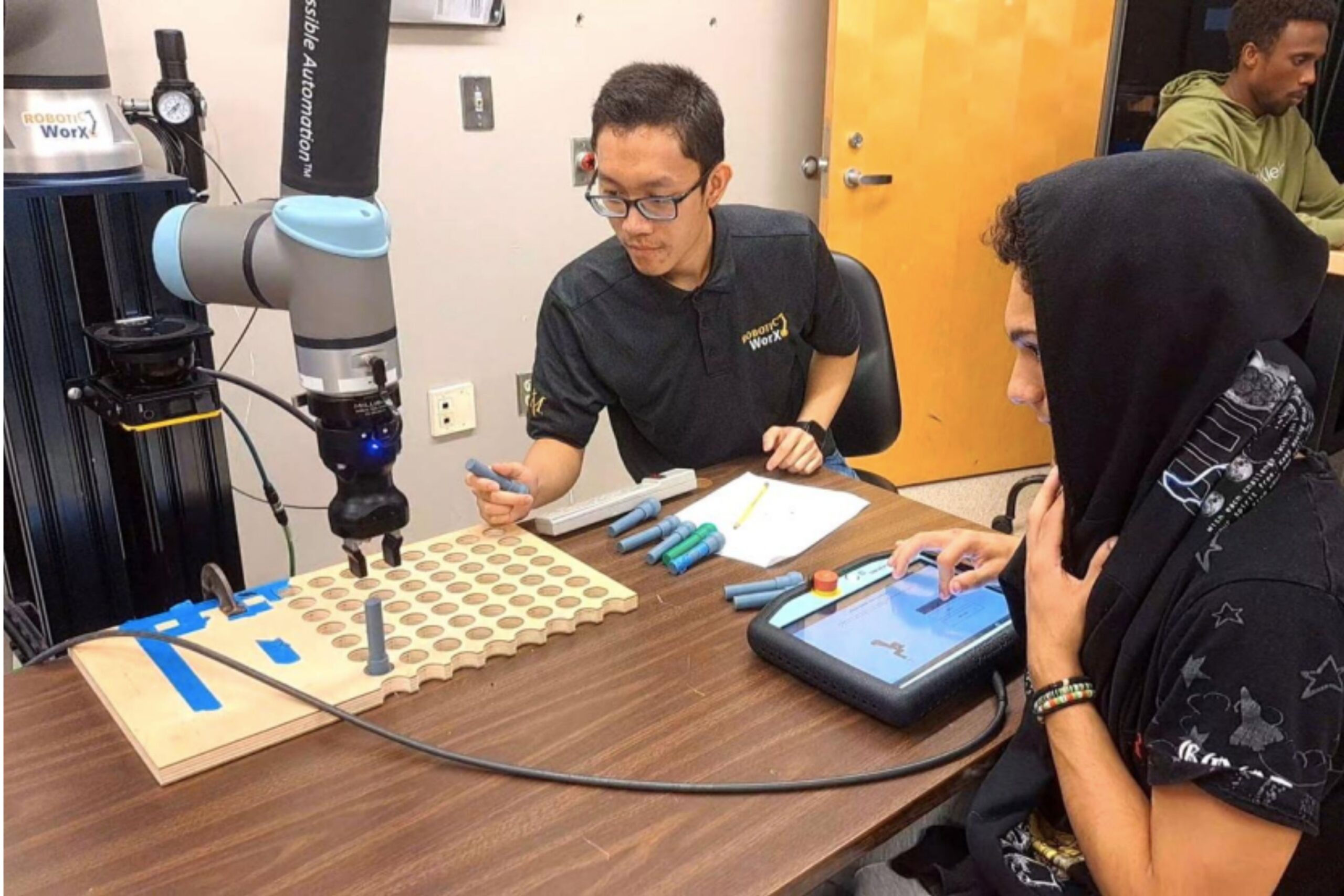 Two people work with a robotic arm to move colored cylinders into a wooden grid, using a control panel in a classroom or lab setting.