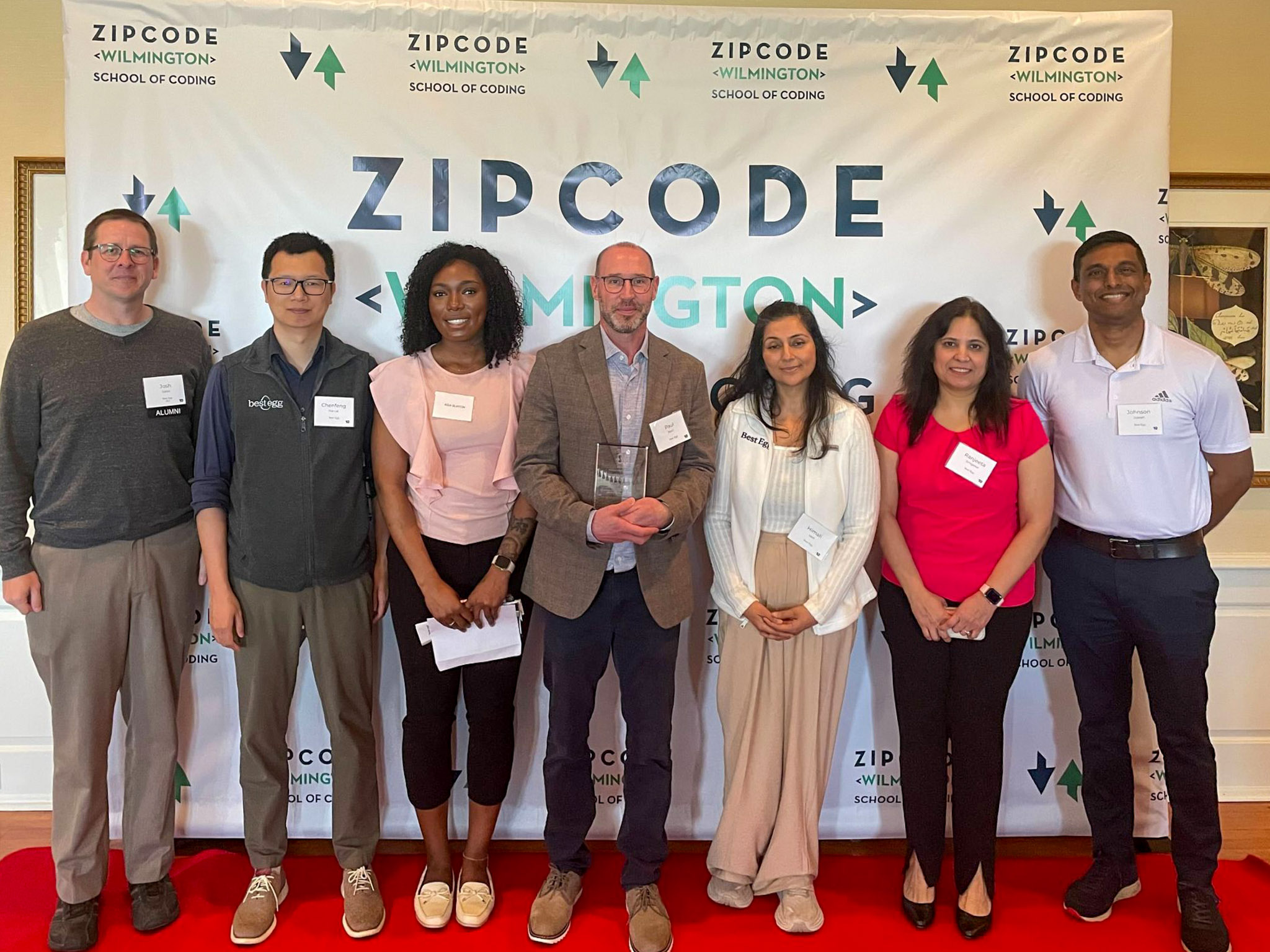 Seven people stand in a row on a red carpet in front of a Zip Code Wilmington School of Coding backdrop, posing for a group photo.