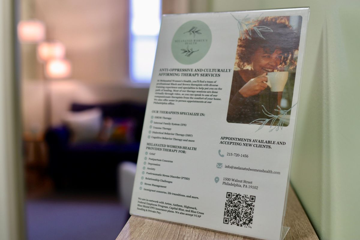 A sign on a table displays information about therapy services at Relational Women’s Health, with contact details and a photo of a smiling woman holding a cup. A blurred couch is in the background.