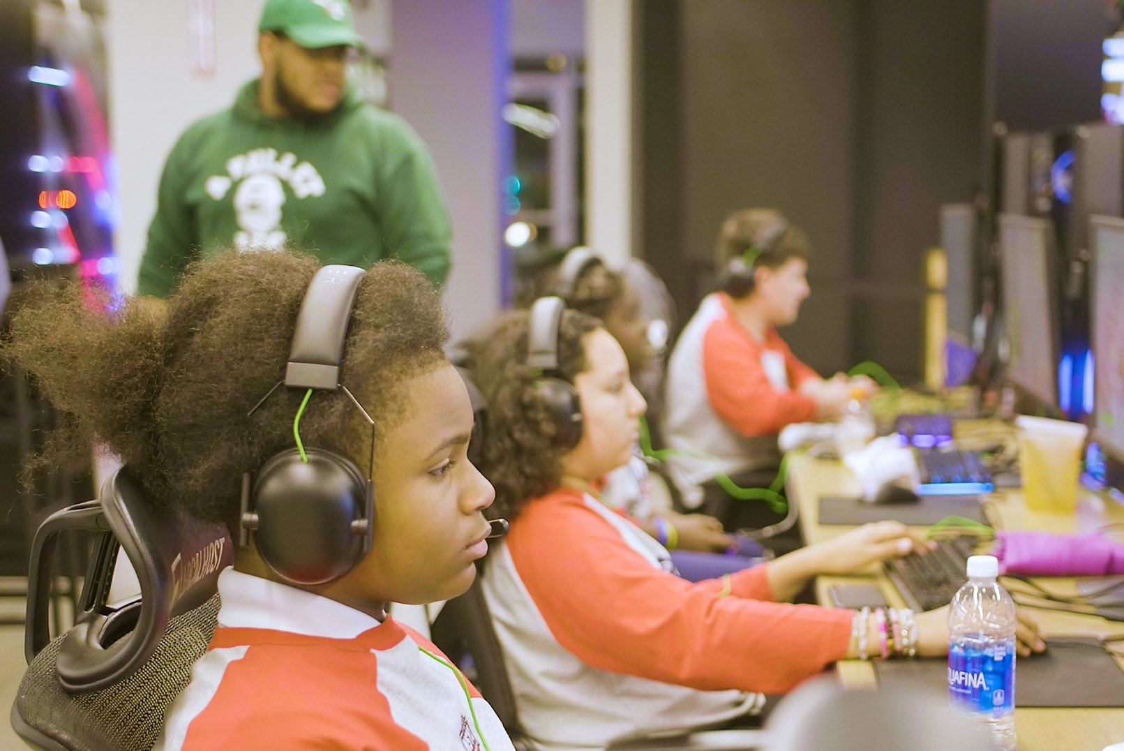 Several young people wearing headsets play video games at computer stations, while a person in a green sweatshirt stands in the background.