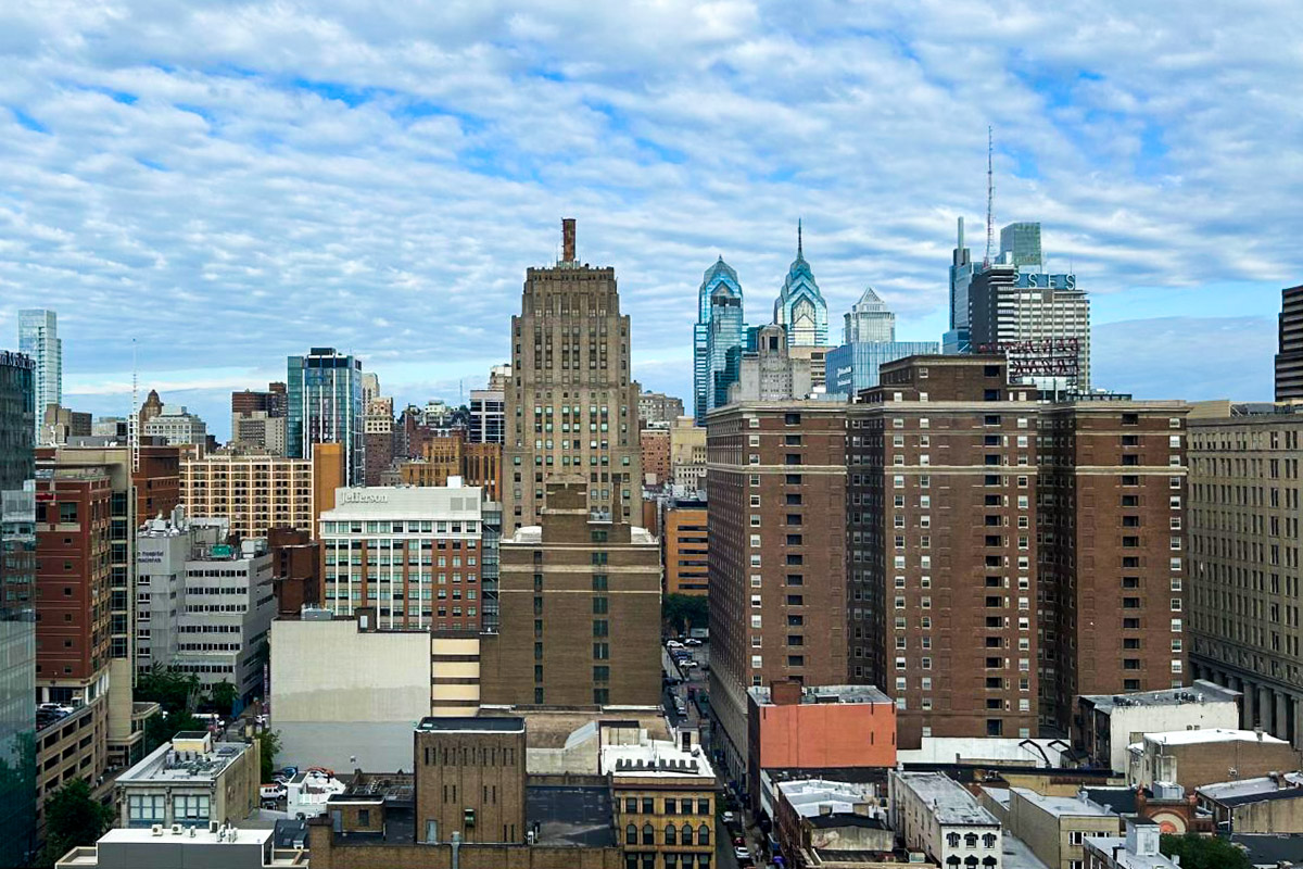 A cityscape view of downtown Philadelphia with tall buildings, including the Liberty Place towers, under a partly cloudy sky.