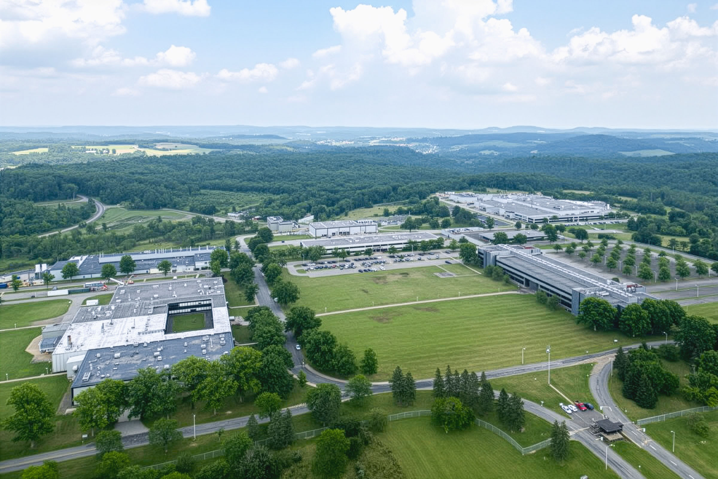 Aerial view of a large office or industrial complex surrounded by green fields, trees, and parking lots, with hills and forests in the background under a partly cloudy sky.