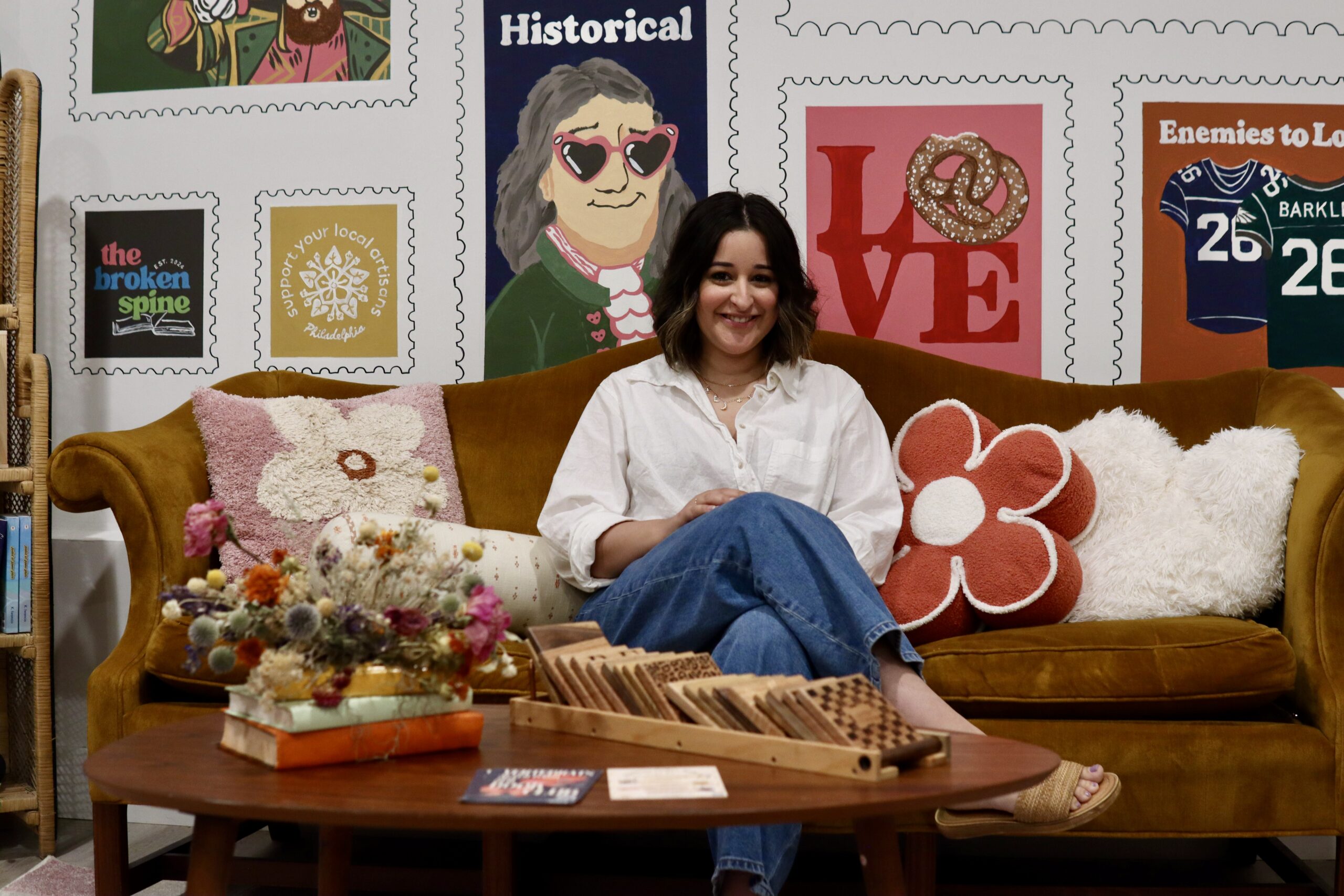 A woman sits on a yellow couch with floral pillows, surrounded by wall art, dried flowers on the table, and wooden puzzles in front of her.