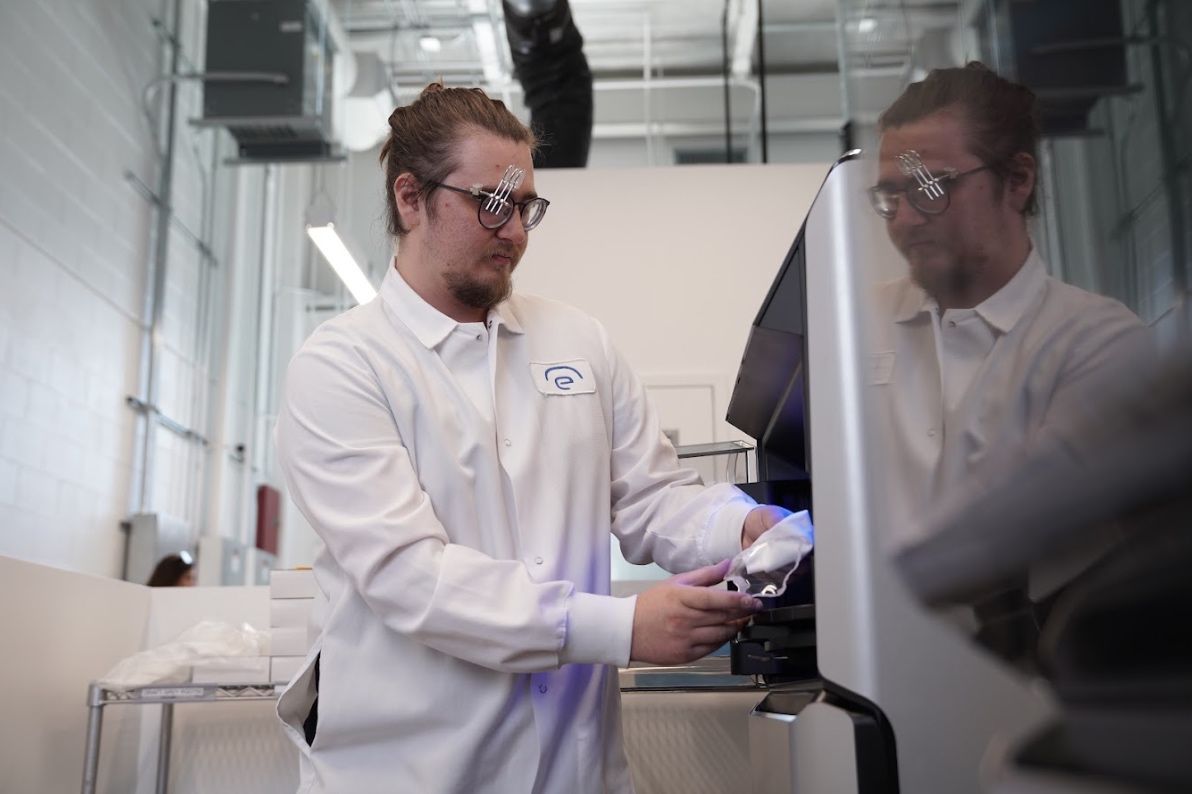 A man in a white lab coat holds a clear device in a lab setting.