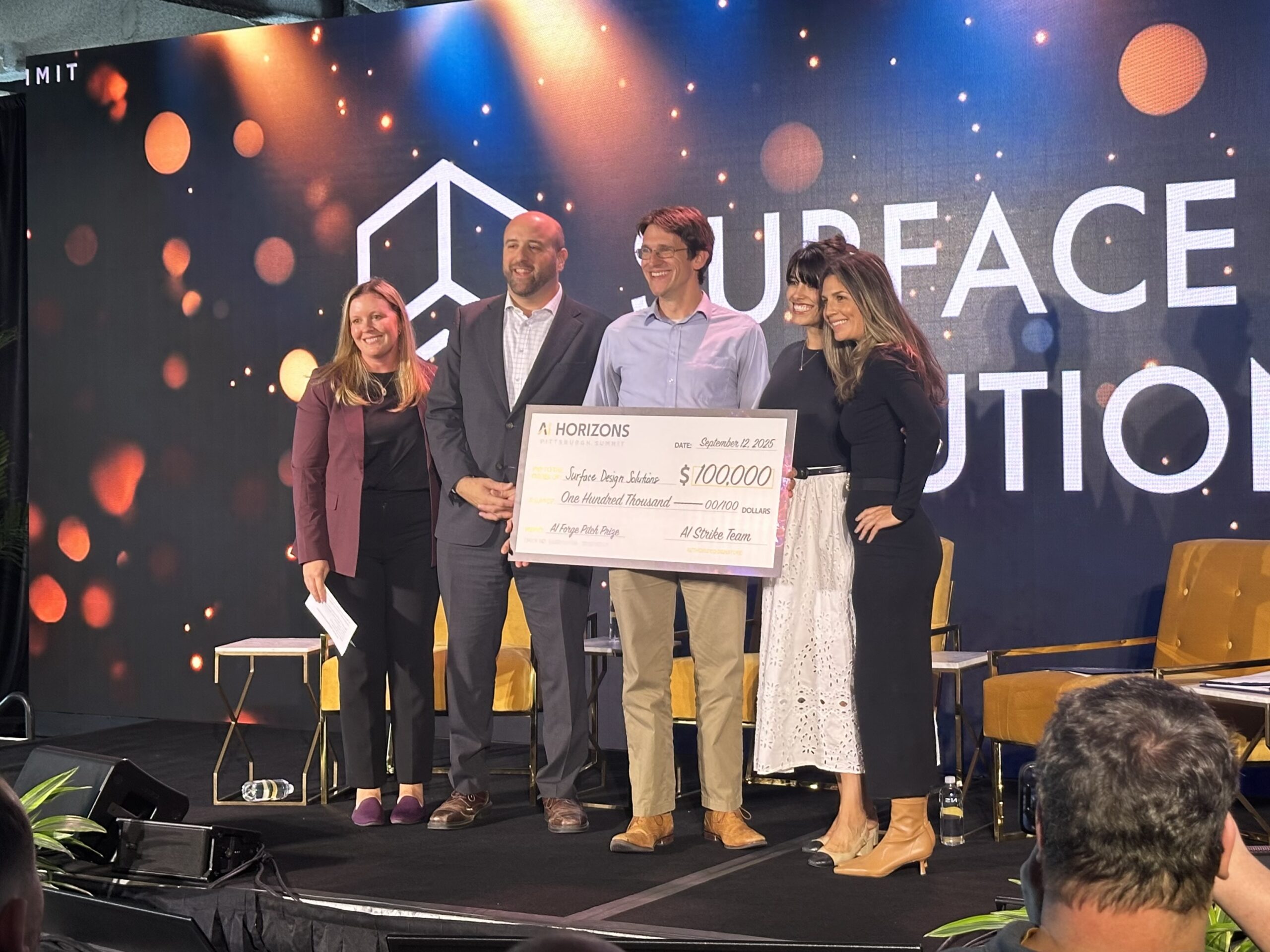 Five people stand on stage holding an oversized check for $100,000 from AI Horizons, with event branding visible in the background.