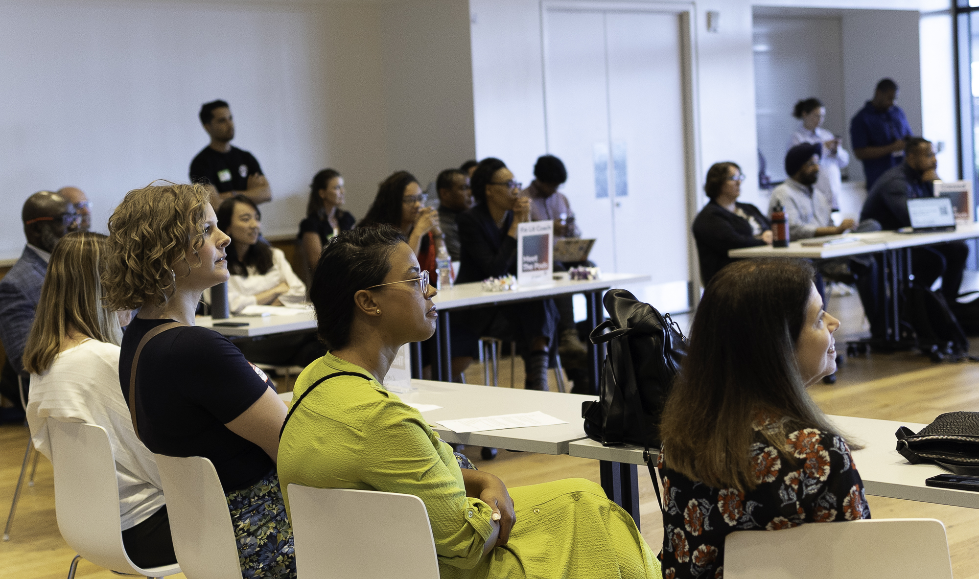 A group of people seated at tables in a bright room, attentively listening to a presentation or speaker out of the frame.