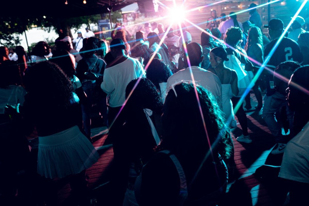 A crowd of people dances at an outdoor event at night, illuminated by colorful lights with a bright lens flare in the background.