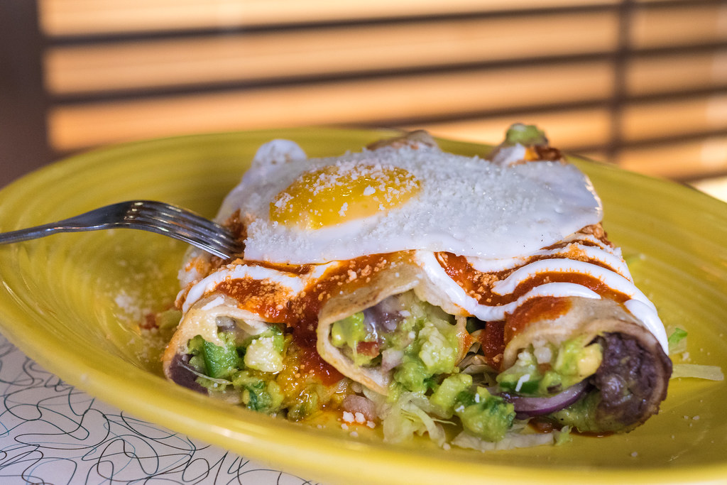 A yellow plate holds three stuffed enchiladas topped with red sauce, crema, grated cheese, and a fried egg, with a fork resting on the dish.