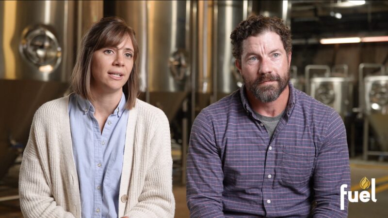 A woman and a man sit side by side in front of large metal tanks, both looking forward. The word "fuel" appears in the lower right corner.