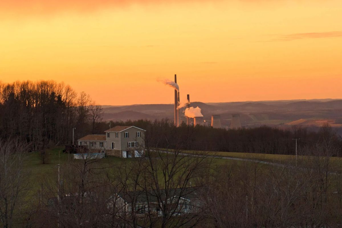 A rural house sits in the foreground with a distant factory emitting smoke under an orange sunset sky.