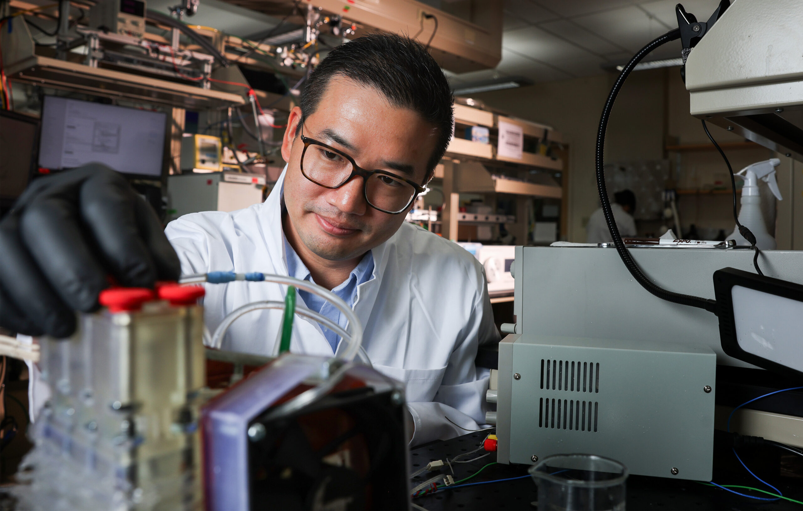 A researcher wearing a lab coat and black gloves adjusts equipment in a laboratory. He is working with wires and tubing connected to a machine.