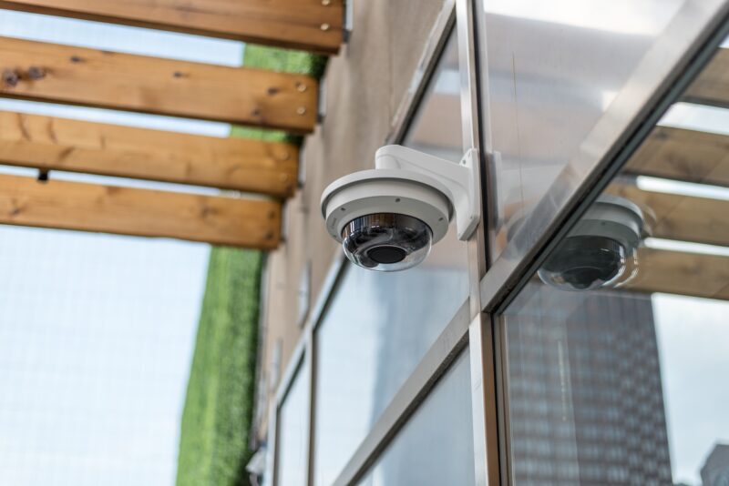 A dome security camera is mounted on the exterior wall of a building near a wooden pergola, reflecting nearby structures in its glass cover.