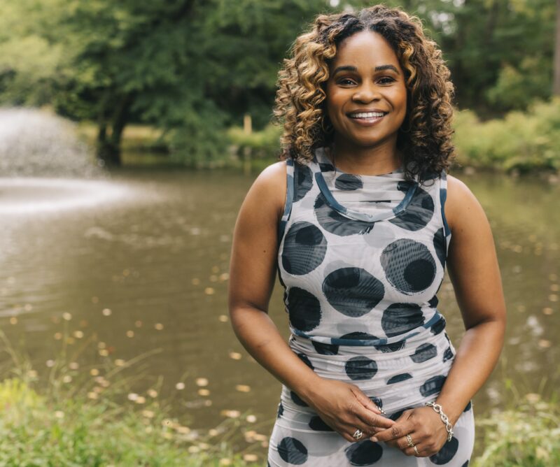 A woman in a sleeveless polka dot dress stands smiling by a pond with a water fountain and trees in the background.