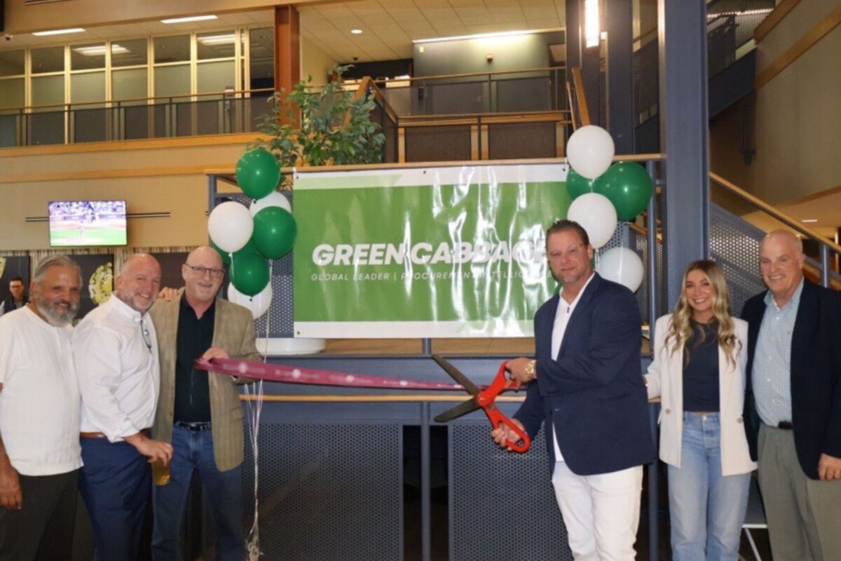 A group of people standing indoors at a ribbon-cutting ceremony in front of a banner with balloons and greenery.