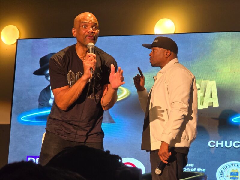 Two men stand on stage speaking into microphones in front of a large screen displaying text and graphics.