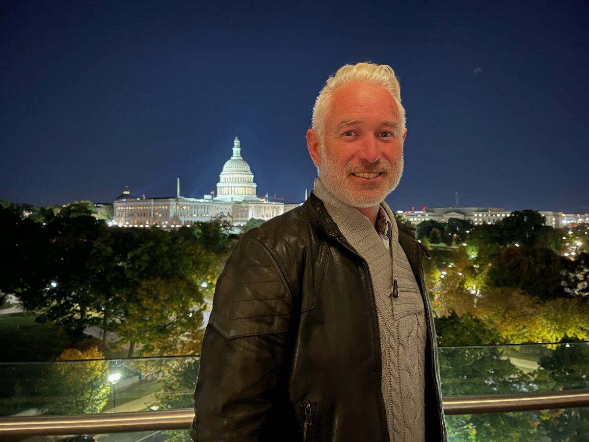 A man with gray hair and a beard stands on a balcony at night, with the illuminated U.S. Capitol building visible in the background.