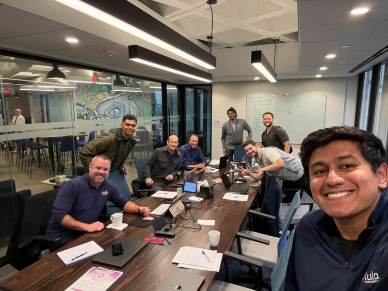 A group of eight people gather around a conference table in a modern office, smiling at the camera during a meeting. Laptops, papers, and coffee cups are on the table.