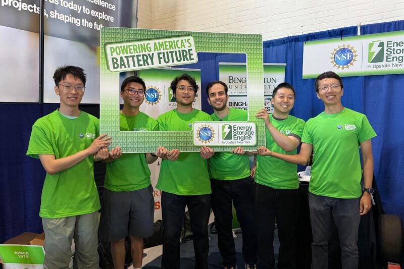 Six people wearing green shirts stand together at an event booth, holding a large frame that reads “Powering America’s Battery Future.” NSF and Energy Storage Engine logos are visible.
