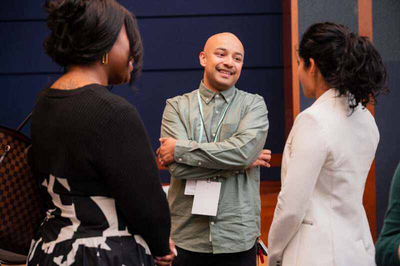 Three people stand in a group having a conversation indoors; one man faces the camera, while two women have their backs turned.