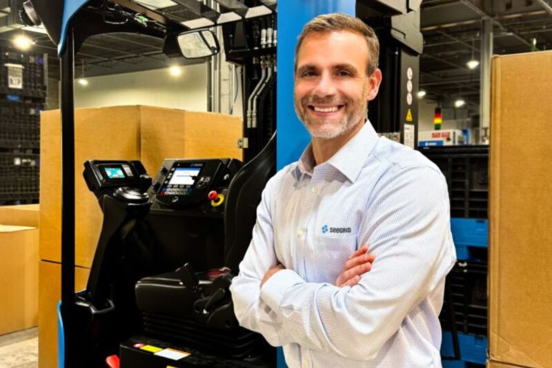 A man in a light-colored shirt stands smiling with arms crossed in front of a forklift in a warehouse setting with boxes and shelving.