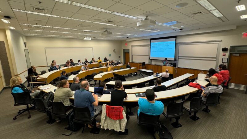 A group of people are seated in a modern lecture hall, listening to a presenter who is speaking at the front and showing a slide on a large screen.