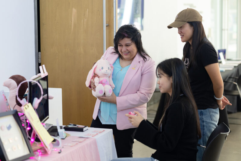 One woman holding a rabbit stuffed animal stands next to two women in black looking at a video game.