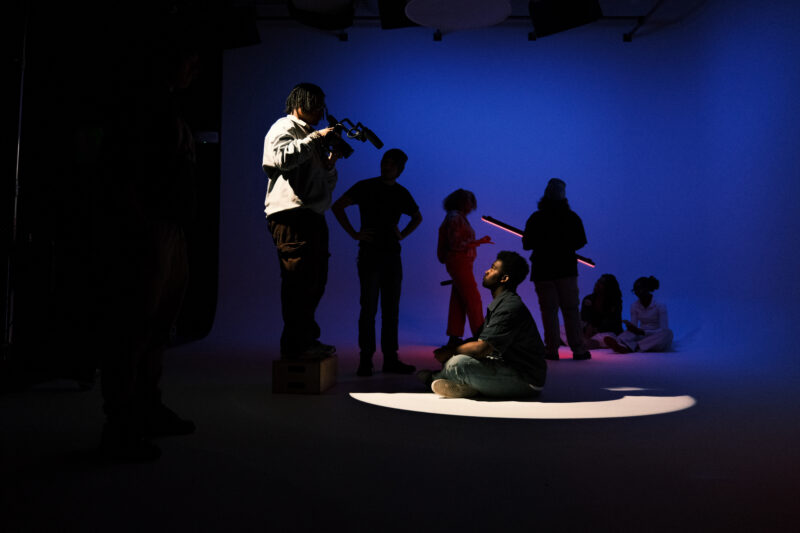 A group of people are on a dimly lit set; one person films another sitting in a spotlight, while several others stand or sit in the background.