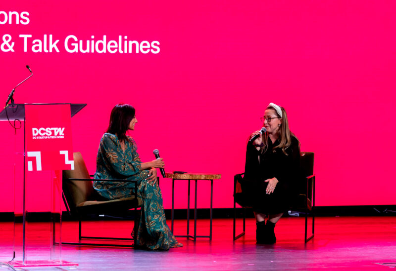 Two women sit on stage with microphones, engaging in conversation in front of a bright pink screen displaying partially visible text.