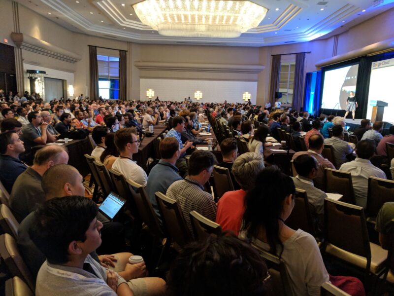 A large audience sits in rows of chairs, attentively listening to a speaker presenting on stage in a well-lit conference room.