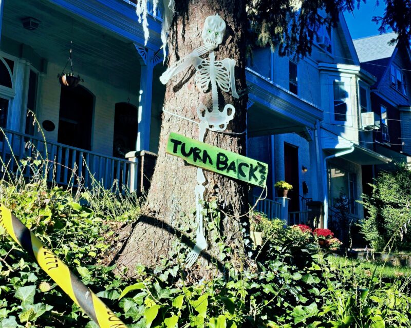 A plastic skeleton is attached to a tree with a green sign reading "TURN BACK" in a yard outside a house, with caution tape in the foreground.