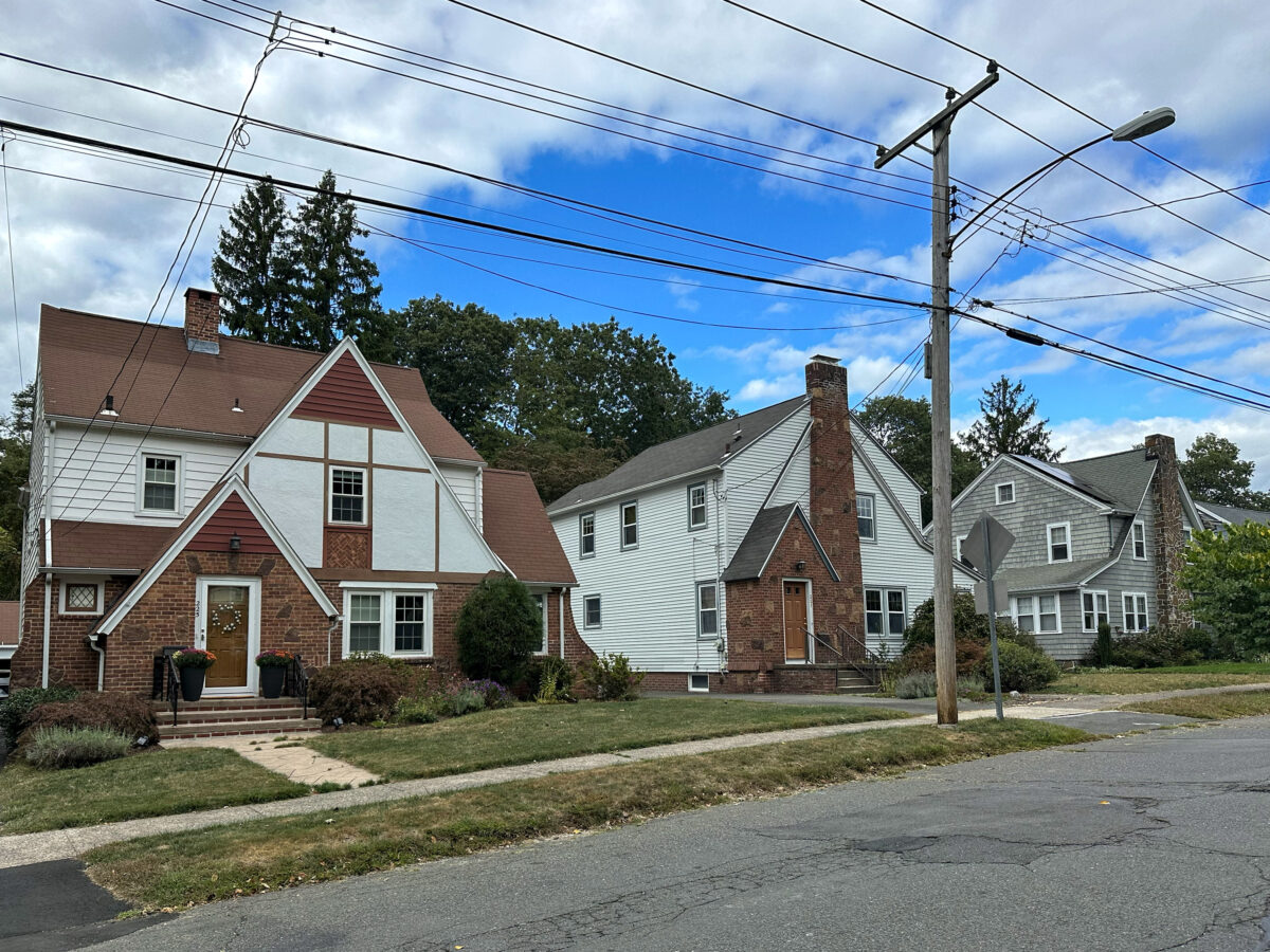 A row of suburban houses with brick and siding exteriors on a quiet street under a partly cloudy sky with visible power lines.