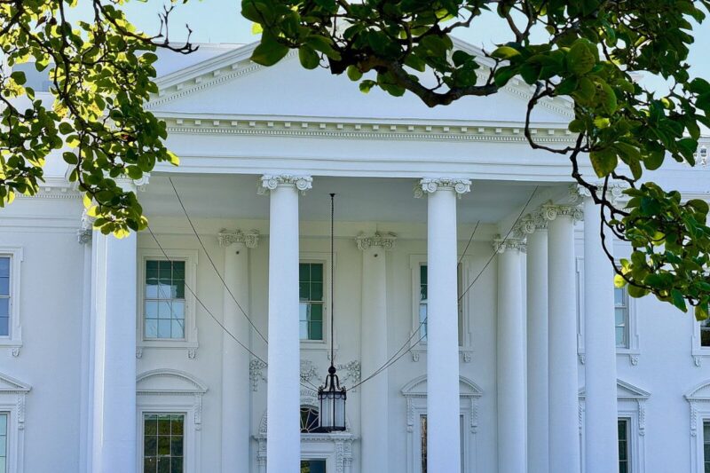 The front facade of the White House with visible columns and decorative trim, partially framed by green tree branches.