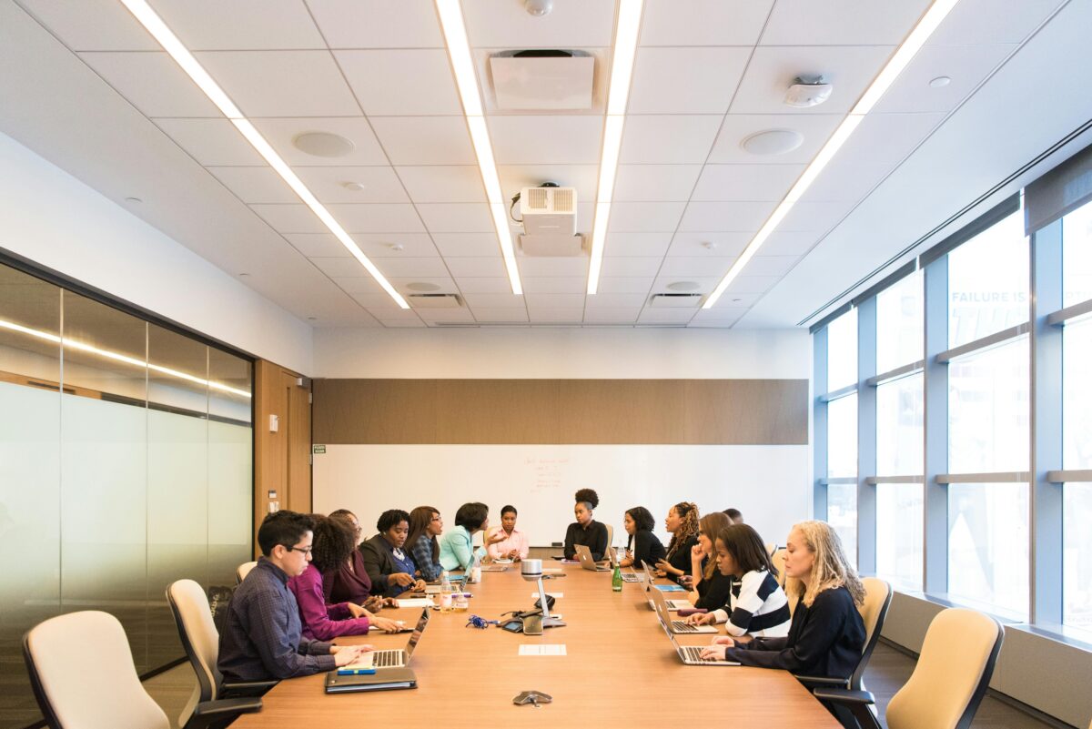 A group of people sit around a long conference table with laptops and documents during a business meeting in a modern office.