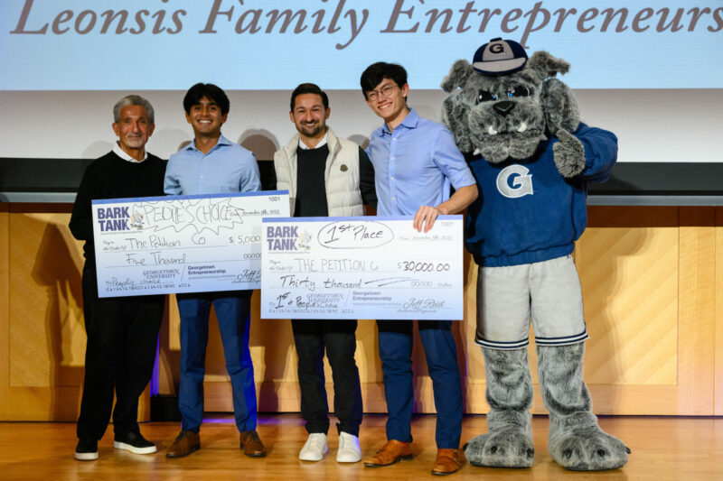 Four men stand on stage holding oversized checks, joined by a person in a bulldog mascot costume. The checks display first and second place awards for a competition called Bark Tank.